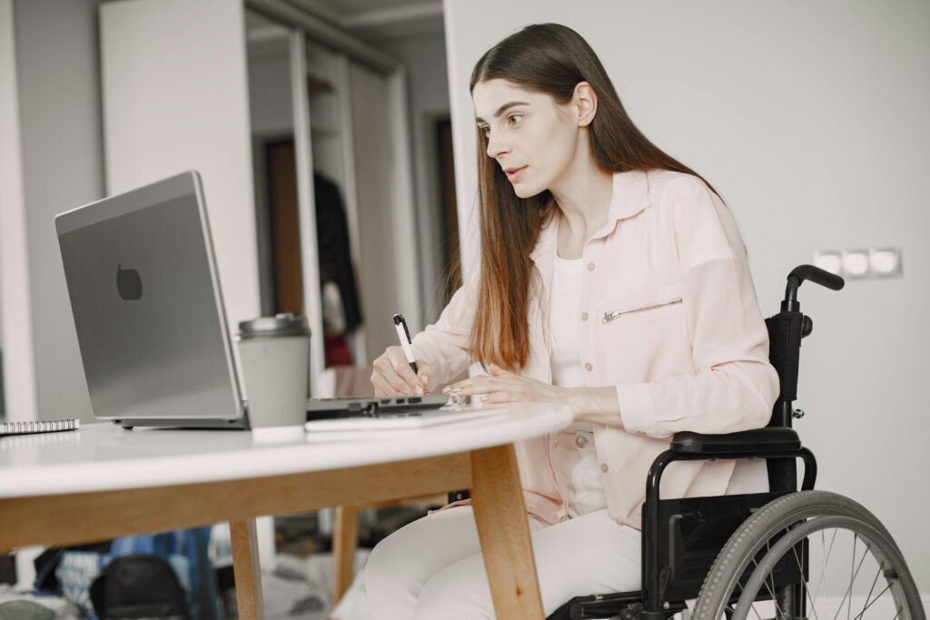 A woman with long brown hair and wearing a light pink top and blazer looks intently at a laptop screen while taking notes with a pen and paper. She is sitting in a wheelchair at a round white and brown wood table.